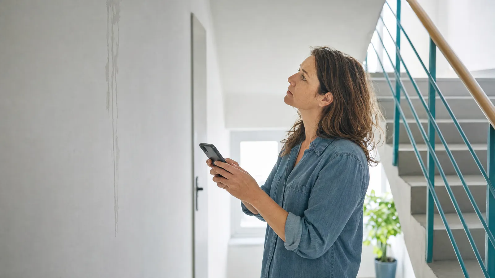 Mieterin im Treppenhaus mit Smartphone in der Hand blickt auf eine Wasserspur an der Wand
